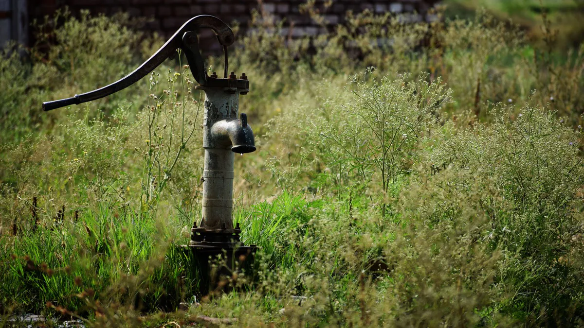 Old manual hand water pump standing in a field of tall grass under open sky