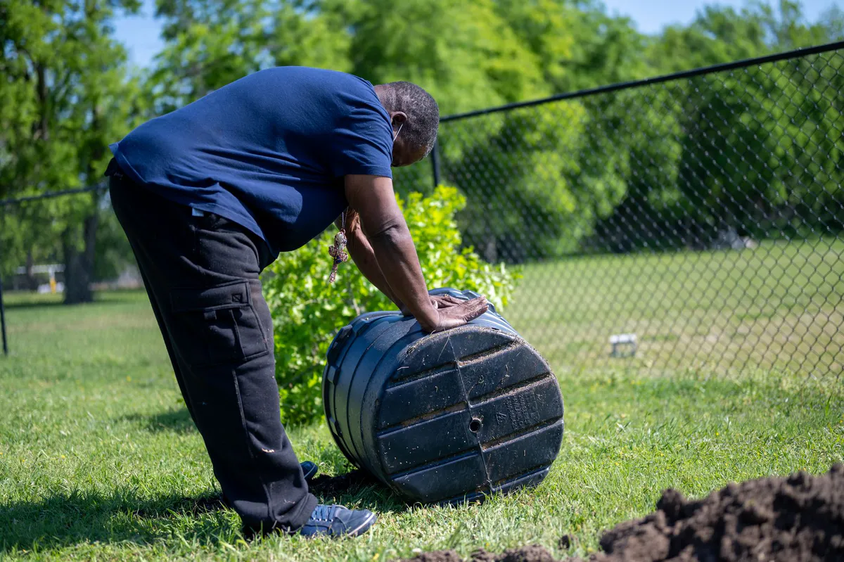 Person tending an enclosed compost bin outdoors on a sunny day in a garden setting