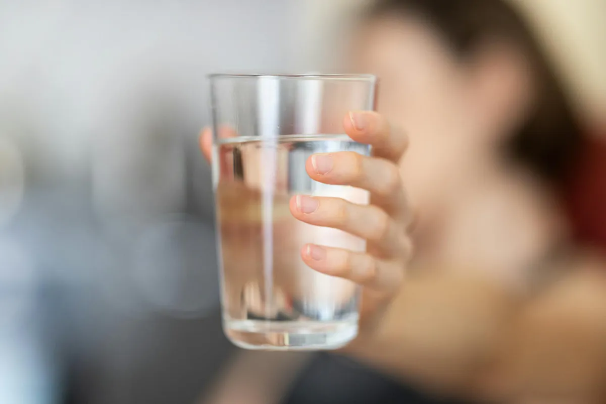 A hand holding a clear glass of drinking water against a soft blurred background